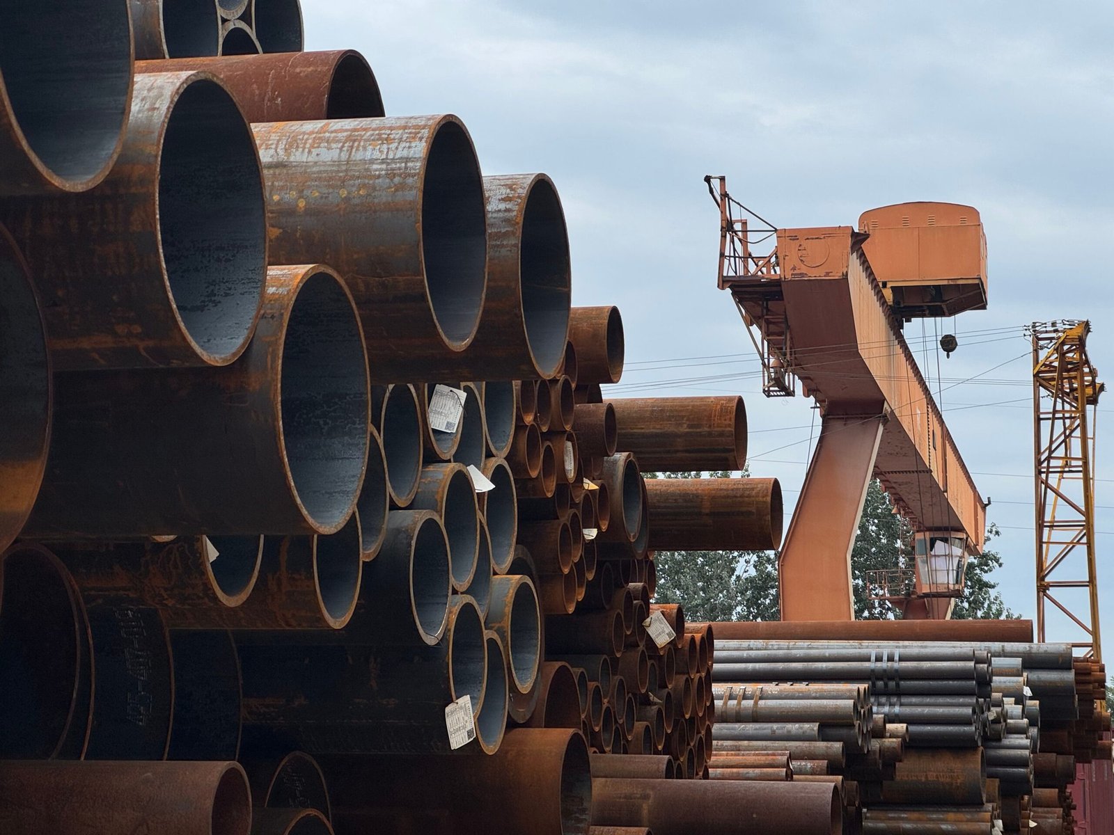 Stacked steel pipes at an industrial yard with an orange gantry crane in the background